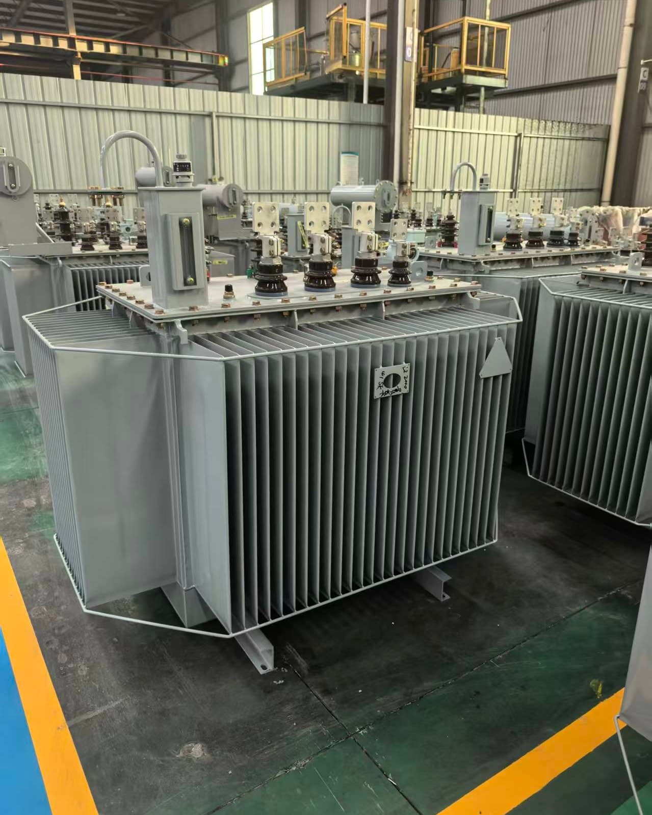 Finished oil-immersed transformers with corrugated tanks, some with conservators, standing in the factory hall